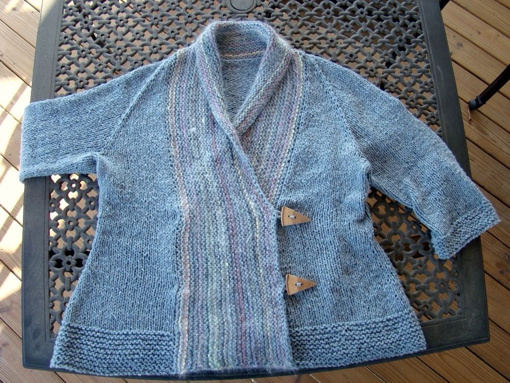 The Iced cardigan lying flat on a black metal deck table with the right sleeve hanging over the edge of the table. The cardigan is buttoned closed, and the shawl collar is slightly folded over around the neck. The striped collar has pastel colors streaked through the blue-grey main color: bits of yellow, green, red, purple, and white. The oversized buttons are pie wedges cut out of a large round branch with the bark still attached, made and sold by The Hickory Tree on Etsy.