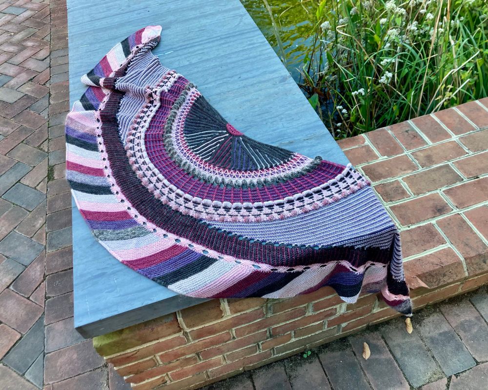 My Shawlography, after its recent brioche-flipping surgery, draped across a grey stone slab and the brickwork under it.  This was Stephen West’s Mystery Knit Along in 2021.  I’m not really a mystery knit along fan—I sometimes knit them after the fact, since I prefer to know what I’m knitting—but I got swept up in this one and it was a blast! The yarn kits from Stephen and Penelope dropped right around my 50th birthday, so I splurged and ordered the Flamingo Cherry Mominoki yarn kit. My pattern modifications were holding a glittery mohair from my stash with the pattern yarn color in a few places (love!), and switching foreground and background colors in the brioche stripe (regret).  Here, I’ve reknit the stripe so the colors are as planned, and the black in the foreground echoes the black in the multicolor fan at the center of the shawl’s crescent.