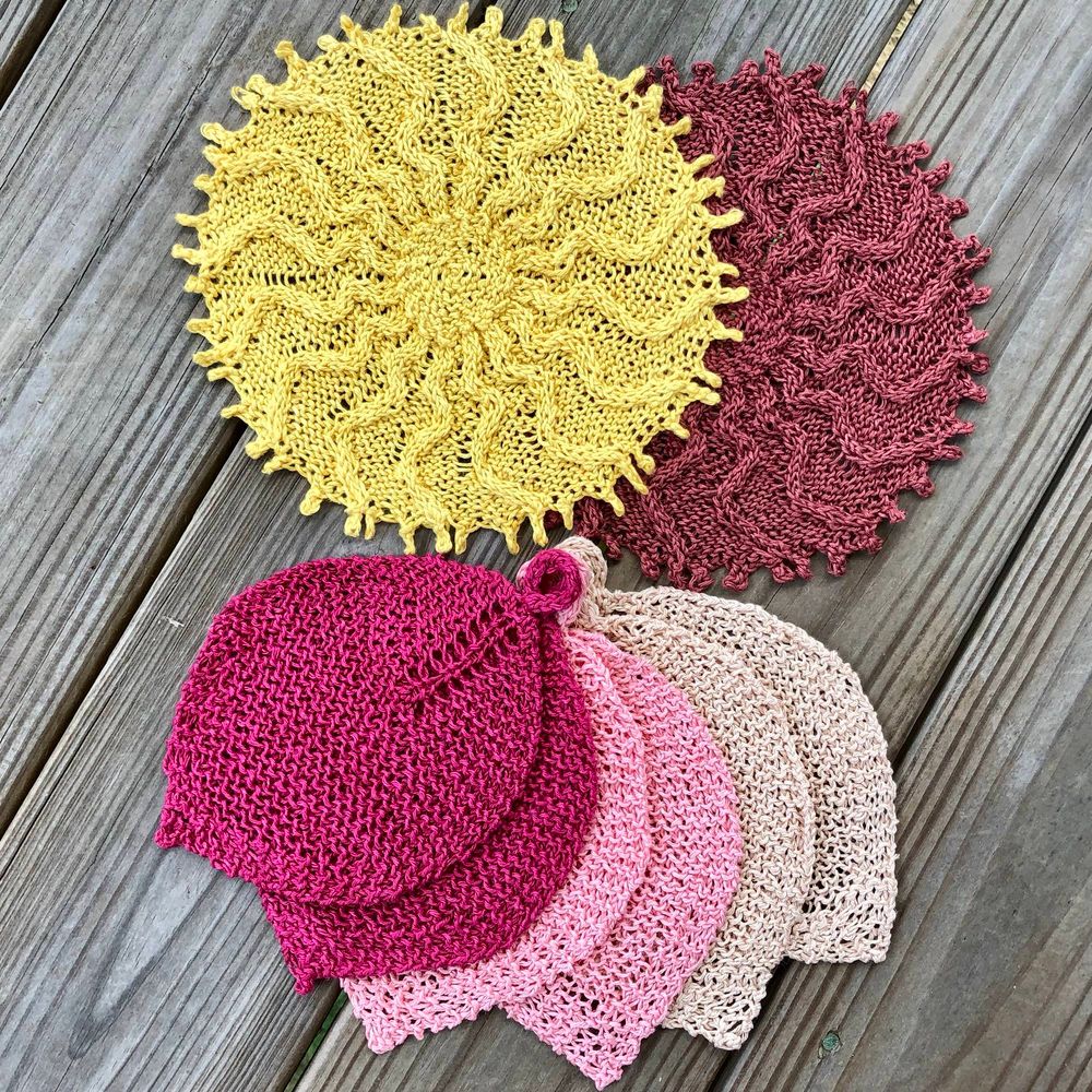 Two Cabled Sunburst Washcloths and three pairs of Bodhi Leaf Washcloths laid on a weathered wooden deck. At the top, a yellow sunburst is half covering a darker sunburst that is somewhere between maroon and beige. Each sunburst is round, with wavy cables radiating outward from a central disk and a bumpy picot edge. At the bottom, the six Bodhi leaves are fanned out. Two are dark pink, two are bright pink, and two are ecru. At the top of each leaf is an i-cord loop for hanging the washcloth.