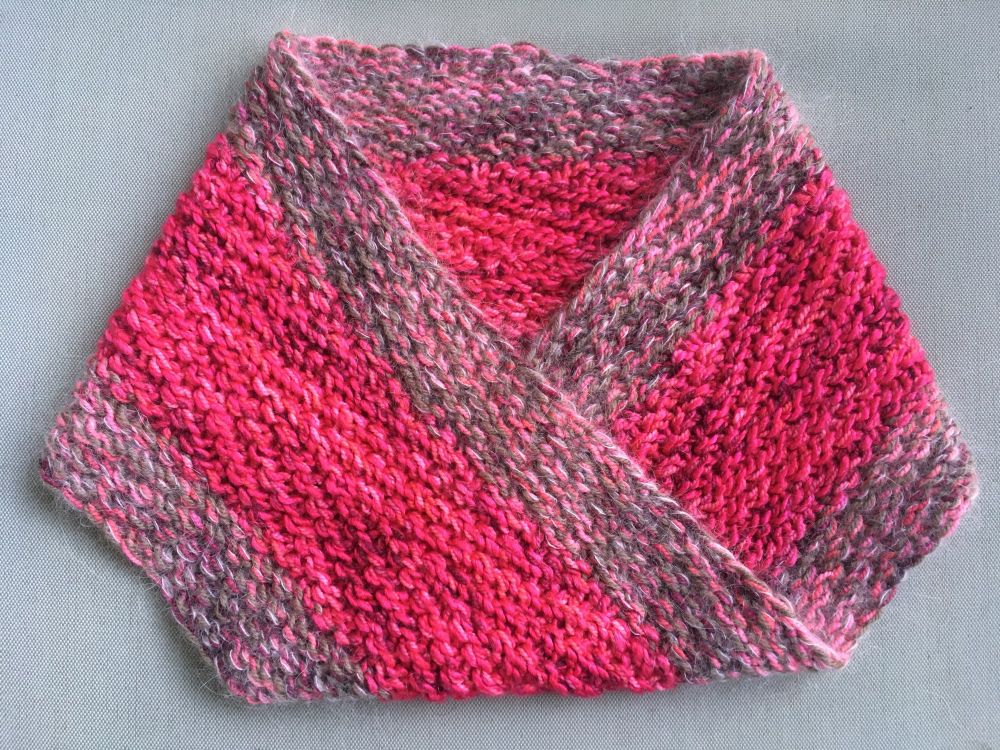 My scrappy Chevron Cowl, made from five lace to sport weight yarns held together in different combinations. The cowl is a seamless möbius band, so it has one surface and one edge. The center of the band is a highly mottled pink, while the border is made from variegated pink and solid brown yarns held together with a pale pink mohair lace. The cowl is folded much as I would put it over my head, with the back horizontal and the front in a downward pointing V. As with möbius cowls in general, it lays in position flatter than a long cylindrical cowl.