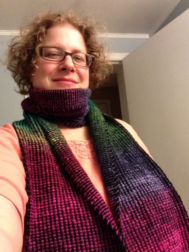 A slightly up-the-nose selfie of a younger me wrapped in the Reversible Stripes scarf she’s just finished. I’m wearing a melon colored top, whose front is mostly covered by the spread-out ends of the scarf running down my chest and off the bottom of the photograph. Both ends are showing the right side, with vertical columns of jewel-toned purl bumps jutting out from the black yarn.  In fact, the black yarn has a similar texture, but being black and slightly thinner, it visually recedes into a backdrop for the brilliant Noro colors. Around my neck and at the inner edges falling from my shoulders, the scarf folds over to show the wrong side. The stripes here are smoother and run horizontally, each stripe a sequence of alternating knitted Vs and yarn slipped to the front.
