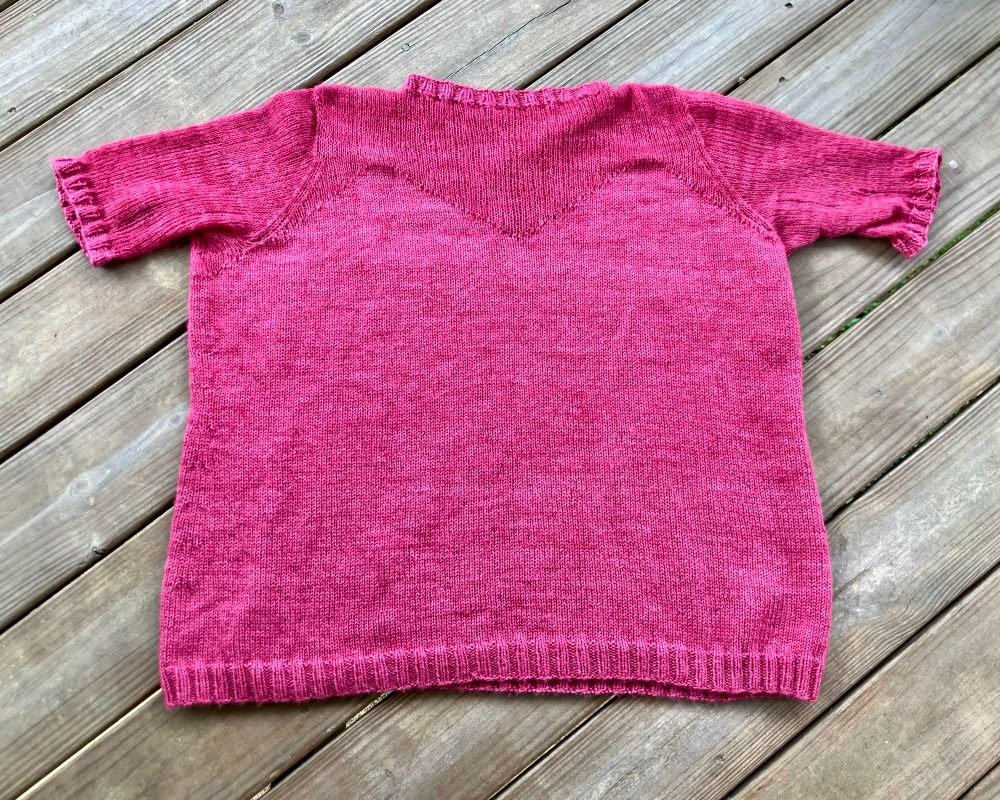 The front of my hot pink As If Lite Tee by Shay Johnson. This rewrite of the As If Tee was a collaboration with Magpie Fibers, and I used their yarns Swanky Sock and Plume. The latter is a cashmere silk blend that looks and feels like a mohair silk lace yarn except softer. The lower body of the sweater and the peaked bust line are knit with both yarns held together, as are the collar and cuffs.  The section above the bust is knit in Plume alone, making it delightfully sheer and airy. This color was part of Magpie’s summer 2023 collection, where all the colors had amazing 70s dance music names.