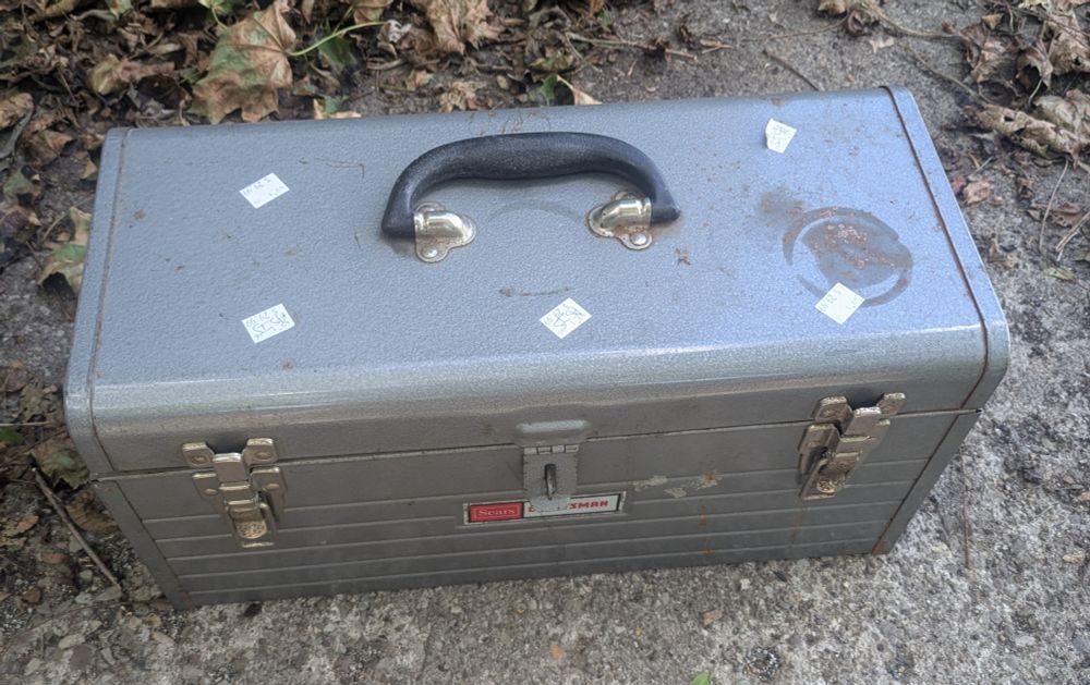 Top, angled view of a grey vintage Sears Craftsman 1970s or earlier metal tool box. It has a gasp for a lock and two latches. There are multiple thrift store stickers with a price of $29.99 on it. 