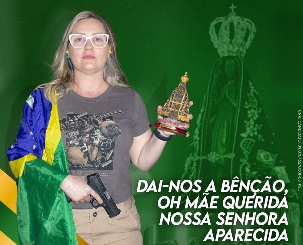 montagem em verde com uma nossa senhora aparecida no fundo e na frente a foto de uma mulher com uma arma na mão direita e nossa senhora aparecida na esquerda, uma bandeira do brasil no ombro e uma blusa onde um buldogue esta com um colete cheio de armas. a mulher tem cabelo loiro curto e usa óculos de grau com armação branca

na imagem esta escrito 

DAI-NOS A BÊNÇÃO, OH MAE QUERIDA NOSSA SENHORA APARECIDA
