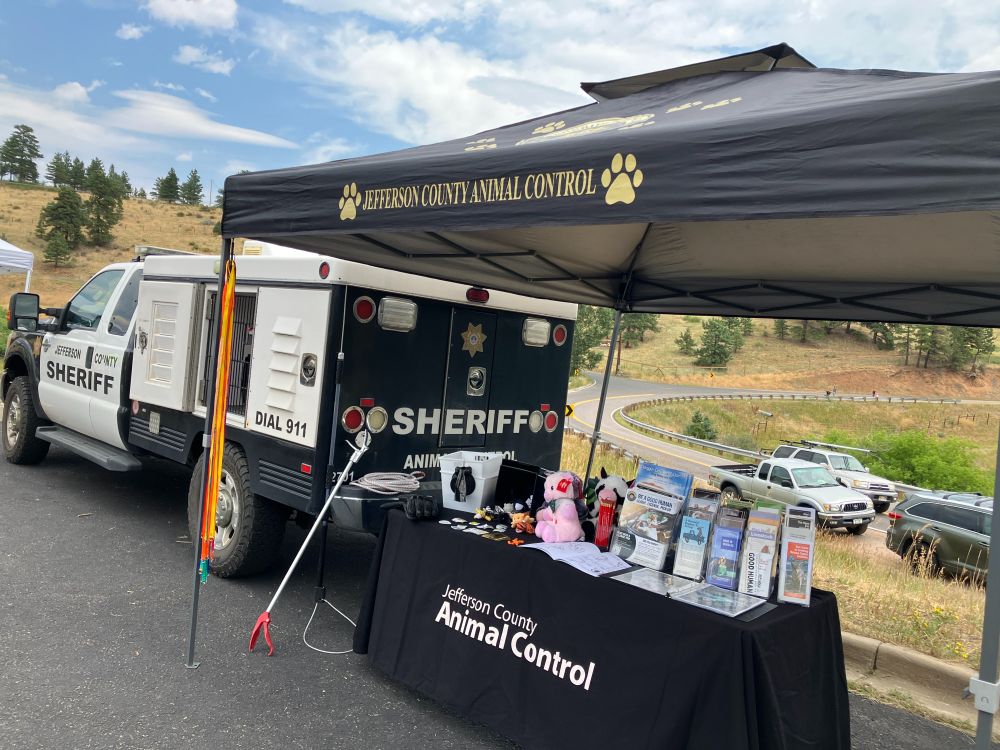 Jefferson County Sheriff Animal Control vehicle parked next to a black canopy tent labeled “Jefferson County Animal Control.” Under the tent is a display table covered with brochures, pamphlets, stuffed animals, and educational materials about animal safety. The scene is outdoors with a curved road, hills, and parked vehicles in the background.