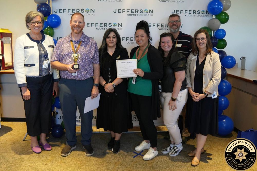 A group of seven people stand indoors in front of a Jefferson County Colorado backdrop, smiling at the camera. One man in a purple patterned shirt holds a gold trophy, and a woman next to him holds a certificate of recognition. The group consists of five women and two men, all in business casual attire. Blue, green, and gray balloons decorate the background.
