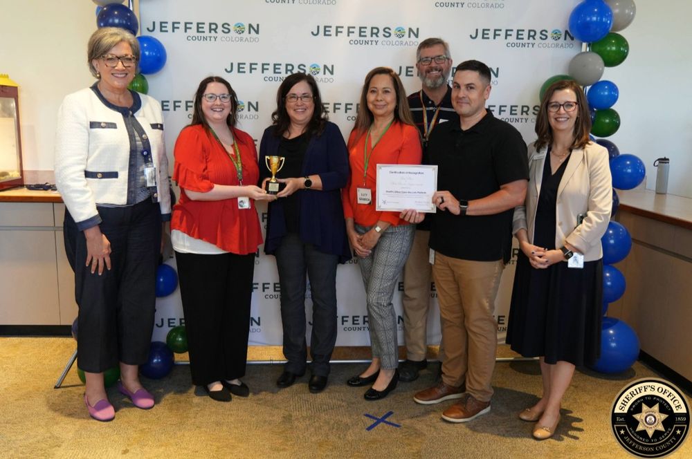 A group of seven people pose indoors in front of a Jefferson County Colorado backdrop, standing on a beige carpet with blue and green balloons along the sides. Two women in the center hold a small gold trophy, and another person holds a certificate that reads 'Certificate of Recognition.' Everyone is smiling at the camera. The group includes four women and three men, dressed in business casual attire.