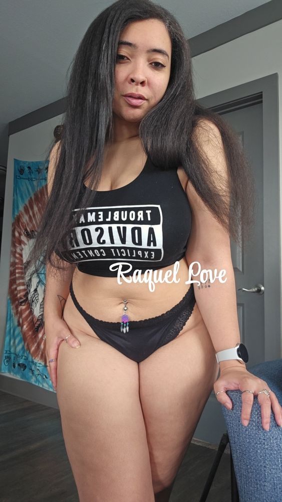Raquel is standing, leaning on a chair with her right hand. She's wearing a black crop top that reads "Troublemaker Advisory Explicit Content" across her breasts. She is also wearing black panties.
