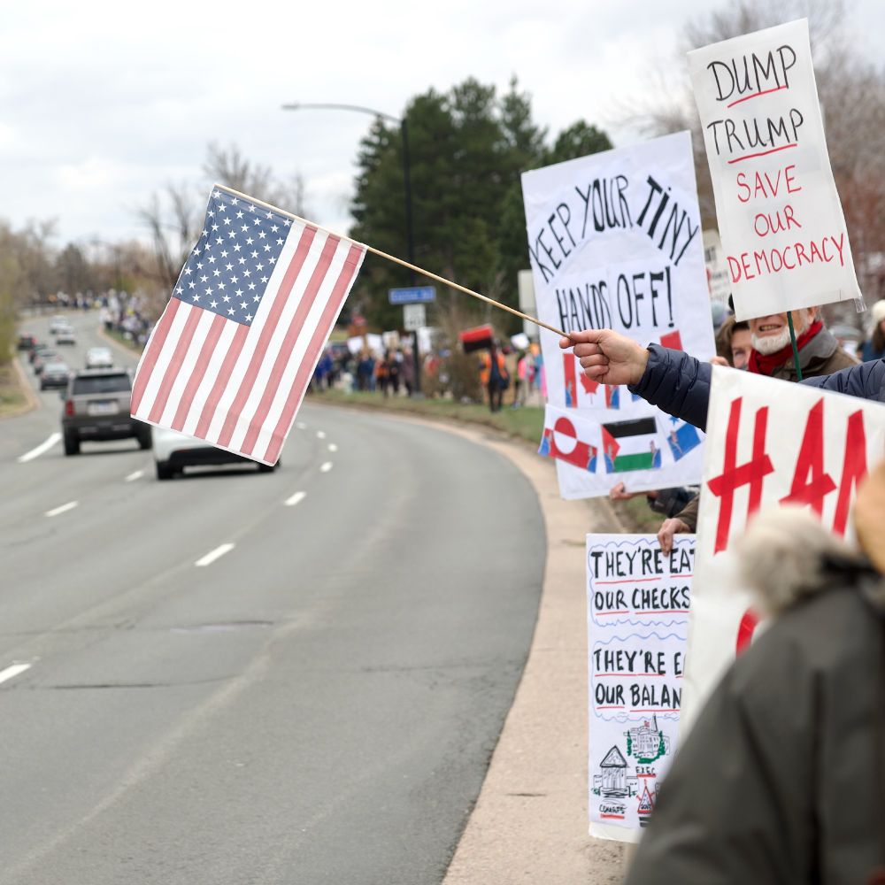 Protesters line the street carrying signs that say "Keep Your Tiny Hands Off!" and "Dump Trump Save Our Democracy!" and waving American Flags