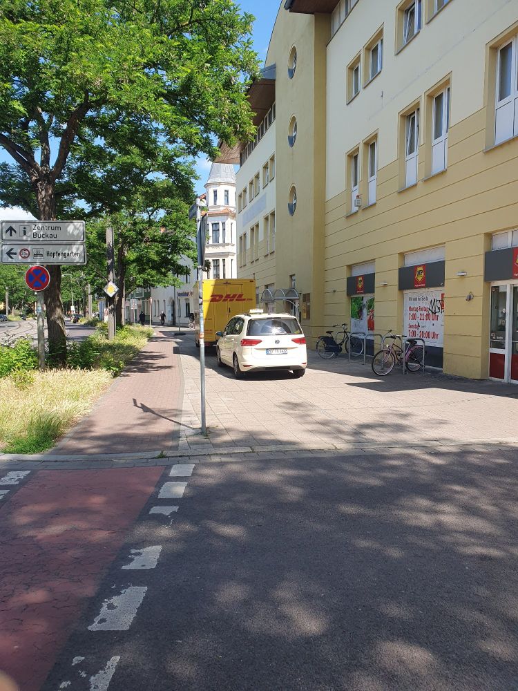 2 Fahrzeuge, ein Taxi und ein Transporter der DHL stehen auf dem Fußweg neben einem Fahrradständer.