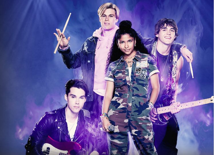 foto promocional da série julie and the phantoms. Julie (adolescente costa riquenha, cabelo cacheado comprido usando um macacão camuflado) está no centro, segurando um microfone. A sua esquerda, agachado, o baixista da banda (garoto branco com topete, jaqueta de couro e um baixo vermelho), atrás dela a esquerda o baterista (garoto loiro alto, segurando baquetas e usando um moletom rosa claro e uma jaqueta jeans preta), ao lado dele O guitarrista e vocalista Luke (o ator Charlie Gillespie, garoto branco de cabelo castanho com corte troy bolton, regata e calça jeans preta, segurando uma guitarra preta com alça branca e vermelha)