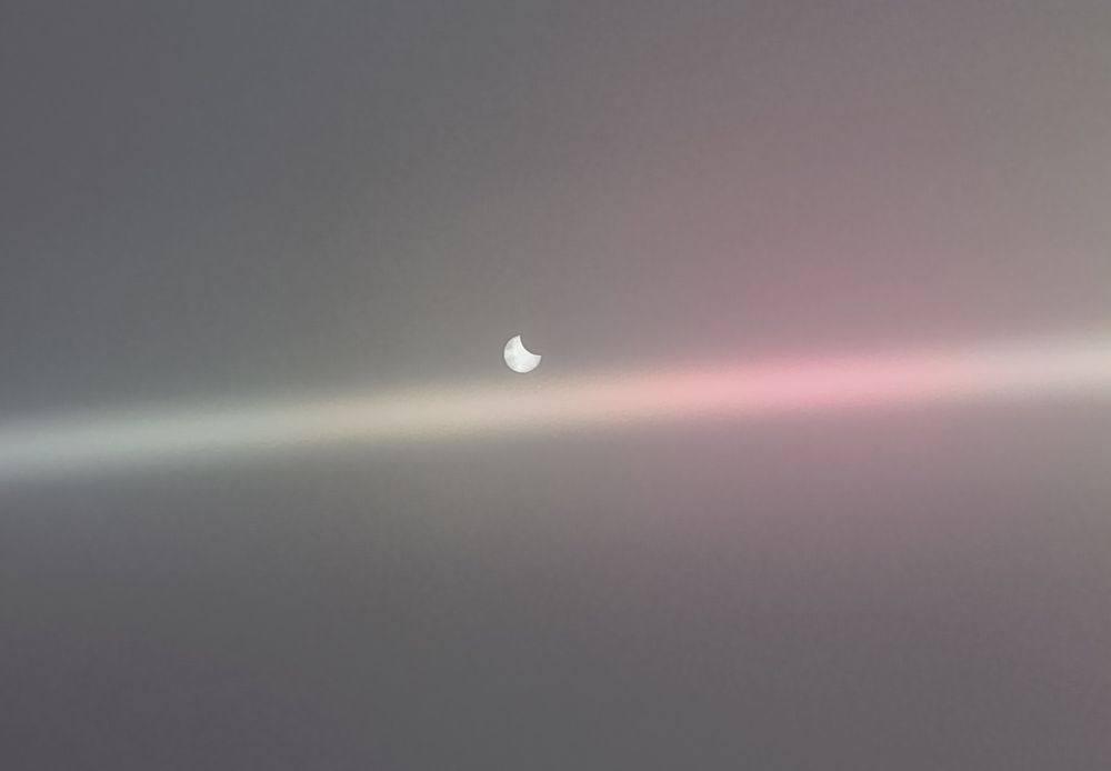 Foto van gedeeltelijke zonsverduistering 29 maart 2025 boven Nederland. Rechtsboven is een hap uit de zon. Gemaakt met mobiel met cd voor de lens, schittering van cd is zichtbaar als een regenboog streep van links naar rechts op de foto.