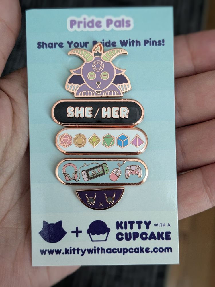 A set of enamel pins clipped vertically to a piece of cardstock. From top to bottom is a Baphomet head, a she/her plaque, a plaque with rainbow-colored polyhedral dice, a plaque with various videogame implements, and a Baphomet butt