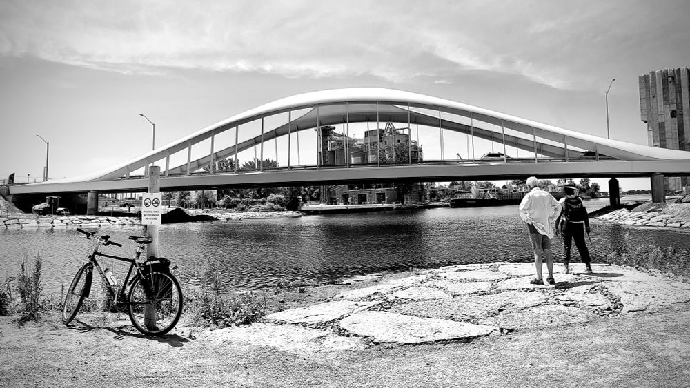 A black and white panoramic photo showing a curving bridge overtop a river. On the left is a bike (my bike!) leaning against a pole. On the right is a couple looking out towards the water. 