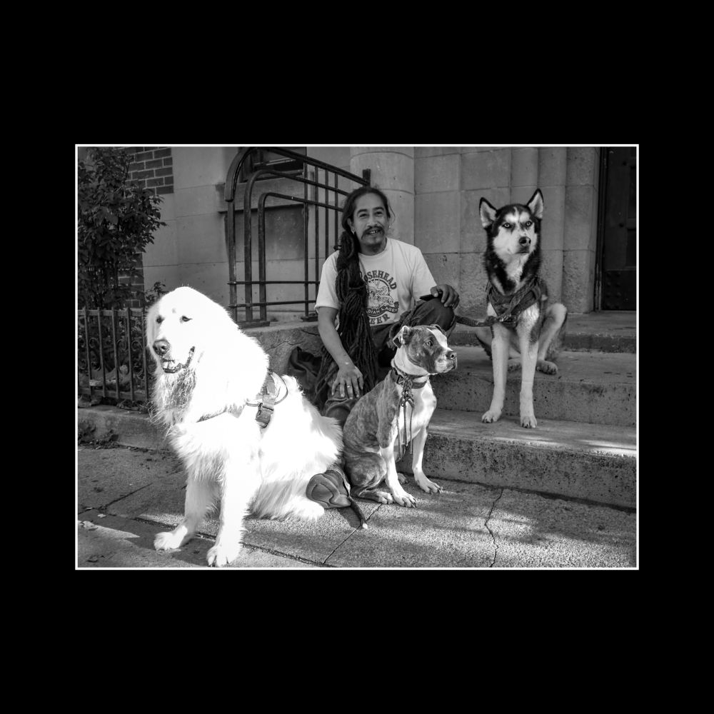 A man named Octavio, with long dark hair and a moustache, sits on a concrete step. His three dogs sit on their haunches in front of him. A large white furred dog names Sosa, a medium sized brown and white dog named Bella and a medium sized black and white dog named Stormy. 