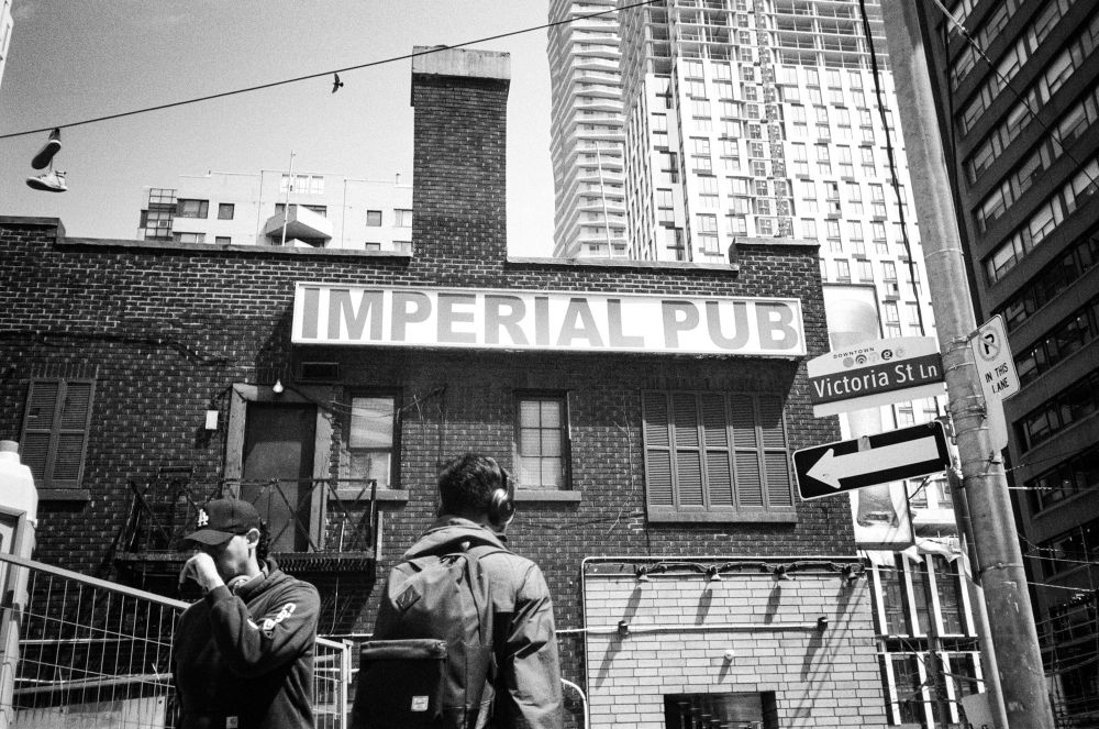 A view looking upwards to a sign on a wall that says “Imperial Pub”. Below are two people walking - one towards the camera and the other away. Above the wall is a power line with a pair of shoes hanging on it. Nearby a bird soars with its wings spread wide.