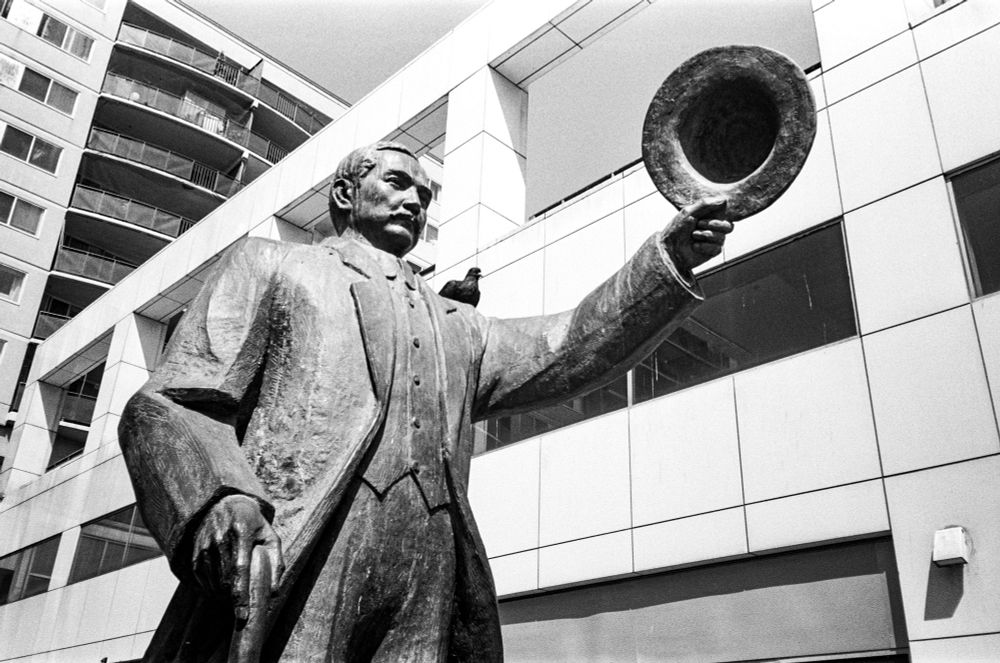 A statue of a man, Dr. Sun Yat-Sen, taken from below. The man is holding out his cap. A pigeon is sitting on his left shoulder.
