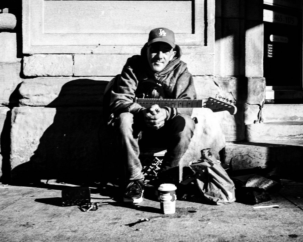 A guitar player sitting on a milk crate with his electric guitar in his lap. He is leaning forward towards the camera, with his hands held together. On the ground in front of him is a coffee cup from Tim Horton’s. 
