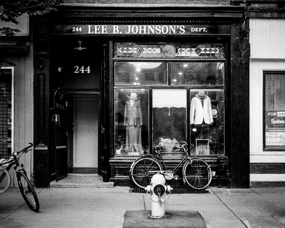 A straight on view of a store named “Lee B. Johnson’s”. A bike leans against the front of the store and a fire hydrant can be seen in front of the bike, in the middle of the sidewalk. 