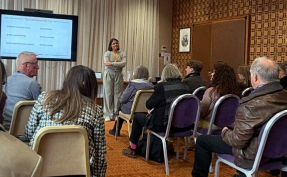 A person presenting to an audience in a conference room with a slide titled "Day-to-Day Communication" visible in the background.