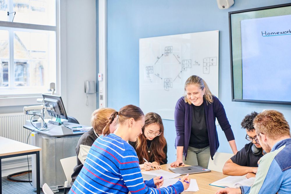 A group of six individuals engaged in a discussion around a table in a classroom. One person is standing, pointing at a document on the table, while the others, seated, look on attentively. A whiteboard with the word "Homework" and a diagram is visible in the background.