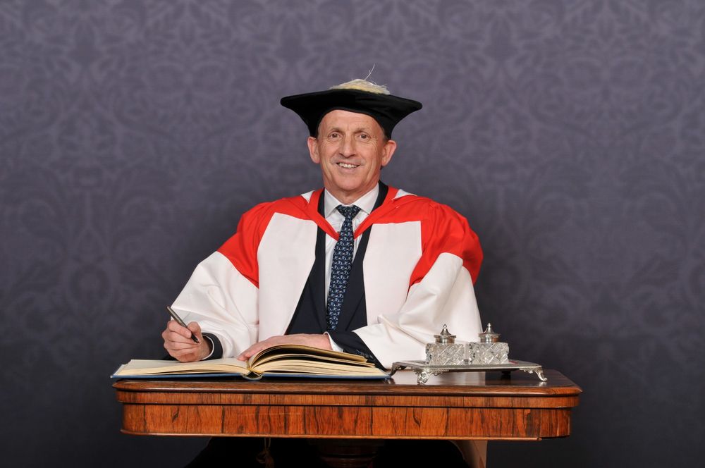 Sir Nigel Wilson in academic regalia, including a mortarboard and a red and white robe, sits at a desk signing a large book. There is a subtle patterned backdrop.