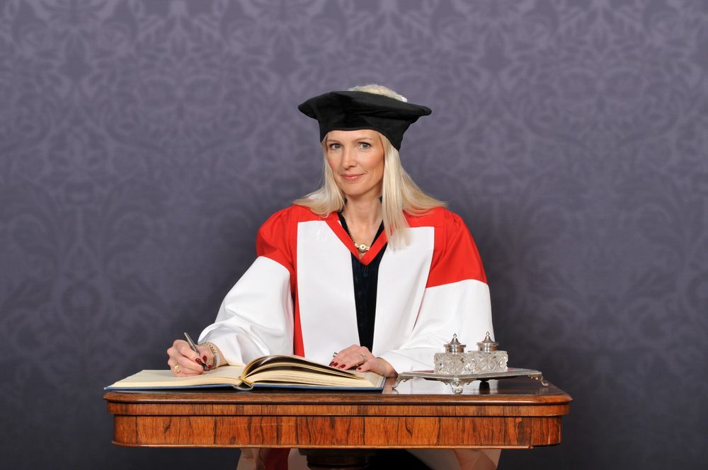 Anna Jones in academic regalia consisting of a black mortarboard, red and white gown, seated at a desk signing a book.