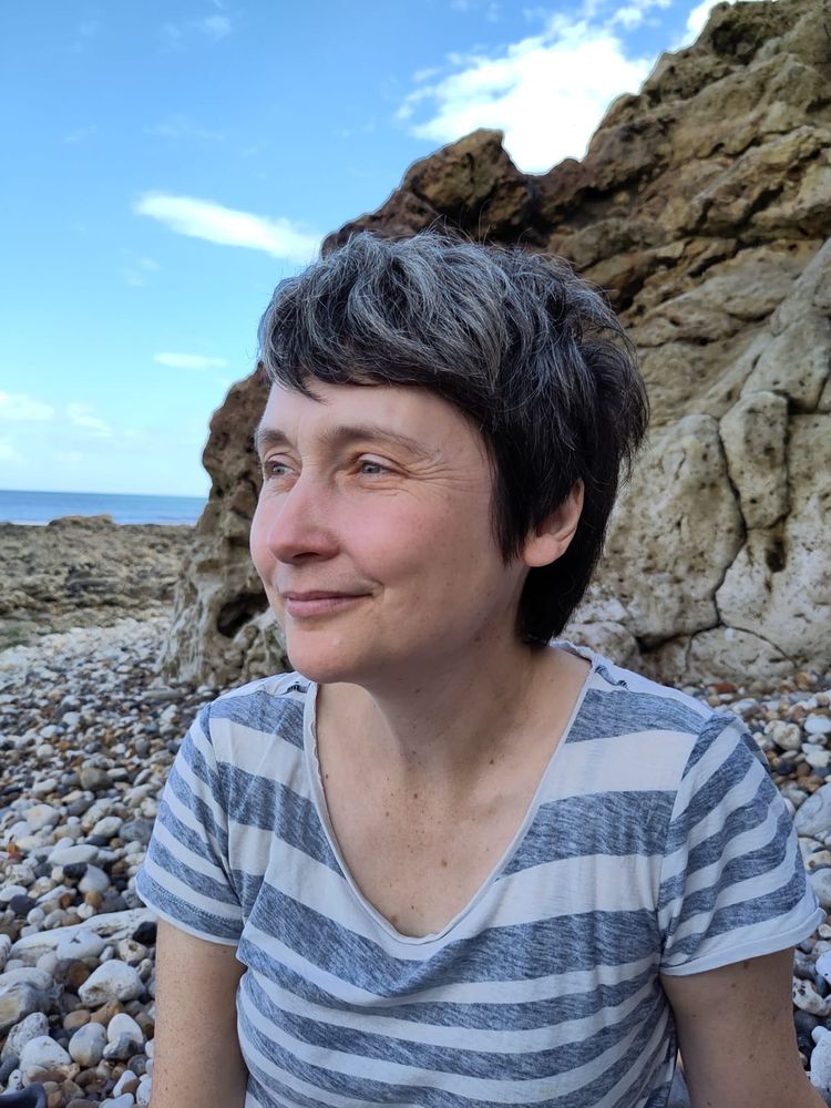 Professor Chantal Conneller sitting on a pebble beach gazing towards the sea, with rocky formations in the background.