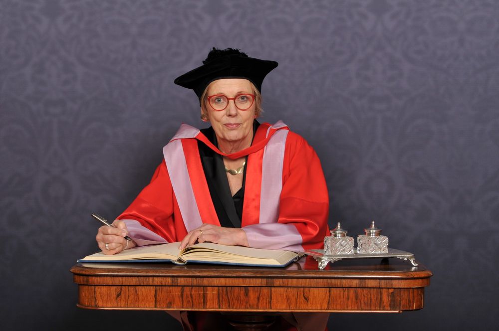 Professor Dame Anne Johnson in academic regalia, including a black mortarboard and red robes, seated at a desk signing a large book. There are two ornate silver inkwells on the desk.