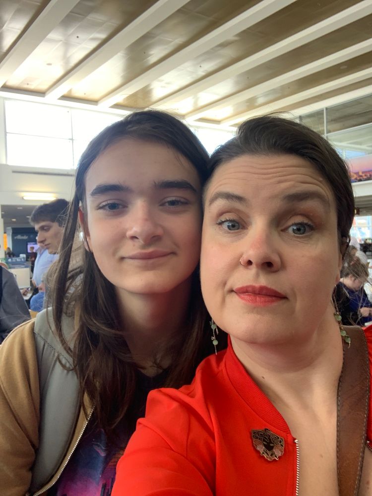White teen boy with long hair cheek to cheek with white middle-aged woman in red lipstick at airport