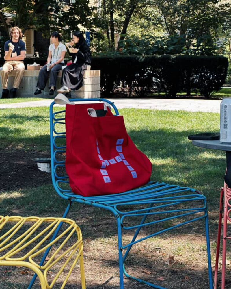 Red tote bag with a white logo placed on a blue wire chair in a park setting with people seated in the background.