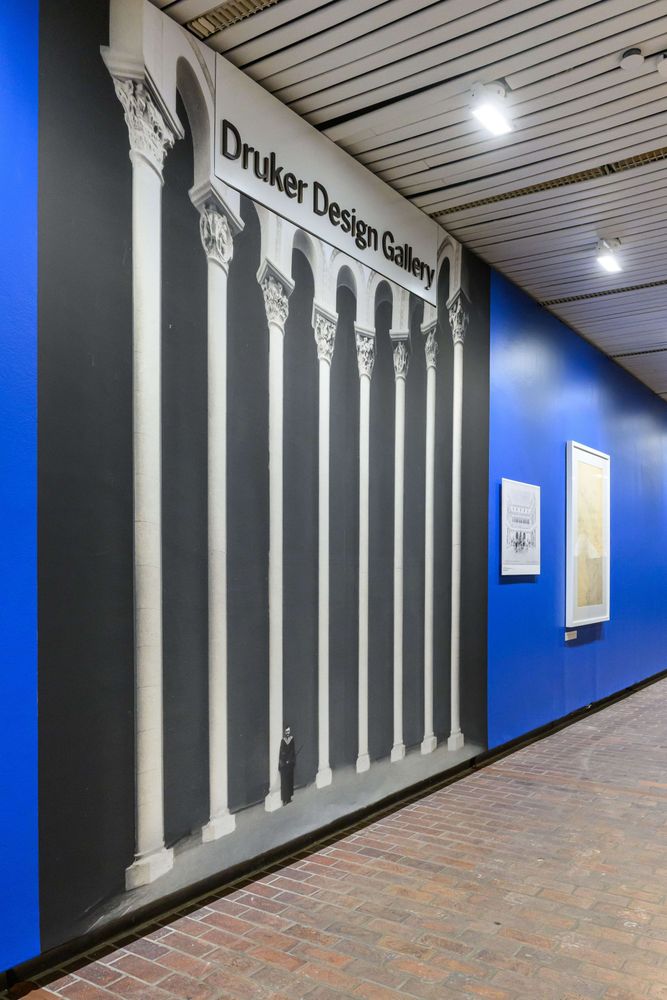The gallery entrance featuring a striking black-and-white mural of classical columns, with additional framed drawings and information panels along a blue wall.
