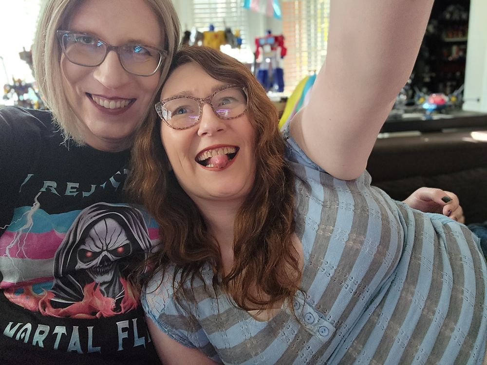 Rachel a blonde in a black trans pride shirt that says "I reject my mortal flesh," taking a selfie while Janey, a Burnette in a striped shirt ALSO, tries to take a selfie.