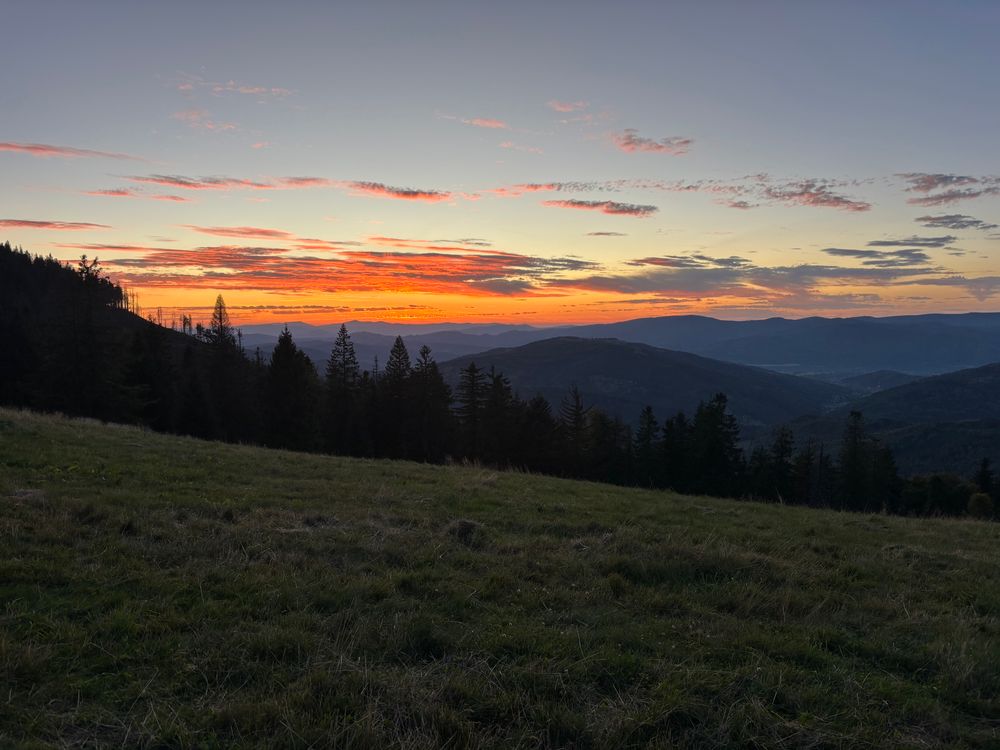 A mountain backdrop seen from a hillside. The sun has gone down, but the clouds are still on fire. It’s autumn, though, so it’s not hot. 