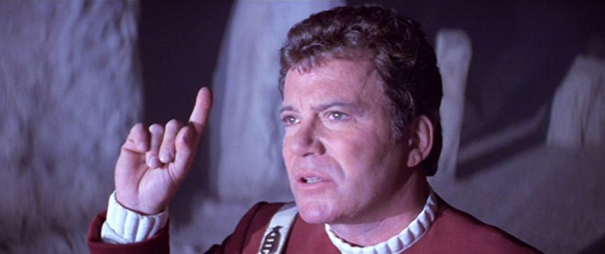 James Kirk standing, bathed in weird light, a questioning expression on his face. He is pointing, upwards.