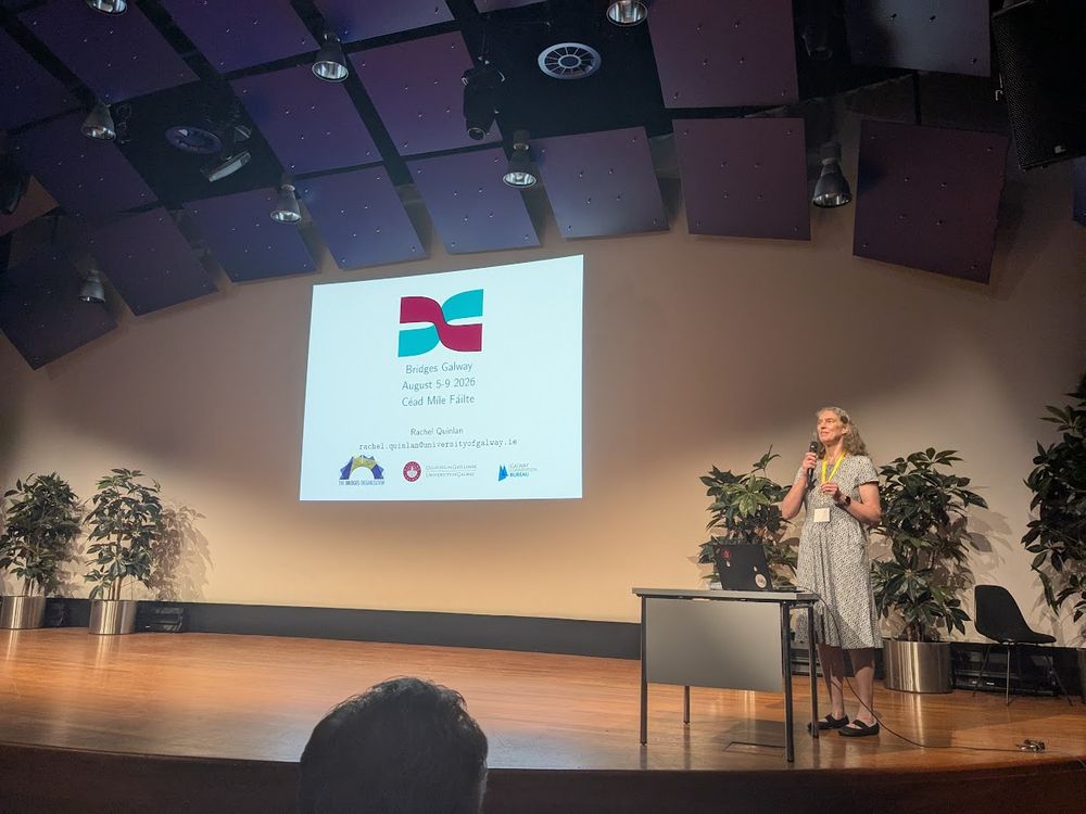 A woman stands on a stage, holding a microphone and speaking, in front of a screen displaying "Bridges Galway August 5-9 2026 Cead Mile Fáilte".