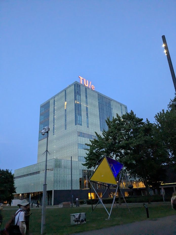 A modern university building, with "Tu/e" lit up on its roof, stands against a twilight sky, accompanied by trees and a colorful geometric sculpture on the campus grounds.