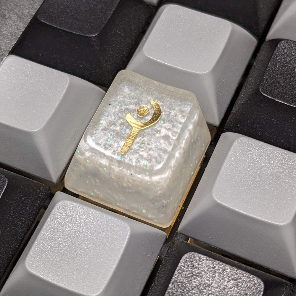 A handmade resin keycap. It has a gold FFXIV White Mage job icon and the base color is a shimmery white. There is some color shifting glitter in the white as well.