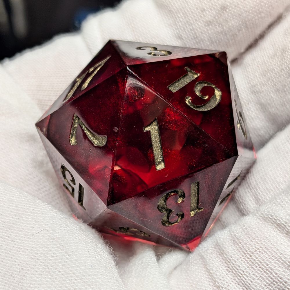 A 30mm handmade resin D20. It is a mixture of shimmery blood red resin and clear resin. The numbers are painted in a dark gold color. In this picture the D20 is being held in a gloved hand and the 3 side is facing upward.