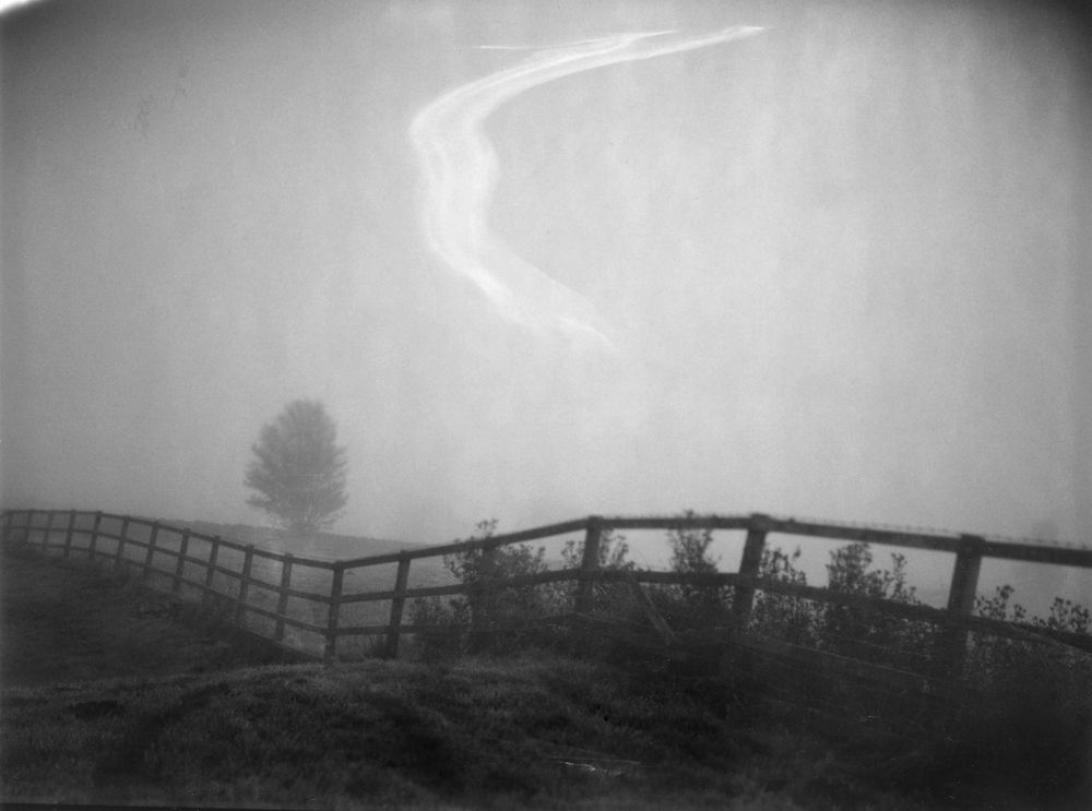 A black and white photograph of a landscape - there is a fence dividing the image with a tree behind it and slightly to the left.  It is a very foggy day and this is reflected in the sky.  There is a streak on the image - looks like smoke rising - the is the out of date film where there are problems with the emulsion.  The image was taken on a 4x5 camera.