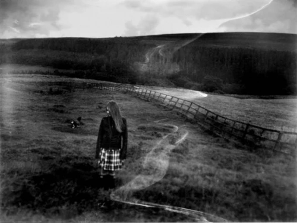A black and white photograph of a girl standing in a field with her dog in the background. There is a white mark, resembling smoke running through the image- this is due to the out of date film that was used. The photo was taken on a large format camera.