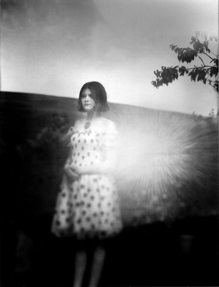 A black and white photograph of my daughter.  The image was taken on a large format camera using out of date instant film - the film has many problems but I also had a problem with light leaks from my bellows - hence the burst of light.  The girl is in a prom dress (she was on her way to a makeshift prom as the original one by the school was cancelled several times due to covid) and standing in the garden - there is a hill behind her.  Her dress looks like polka dot but is actually full of strawberries.  There is a burst of light to the right hand side and a tree encroaching just above.