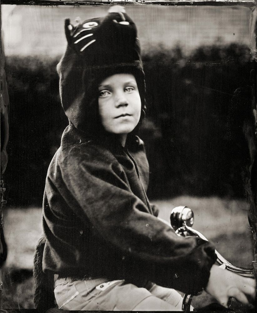 A wet plate collodion portrait of a child dressed in a cat costume - she is ride a tricycle and staring directly into the camera.