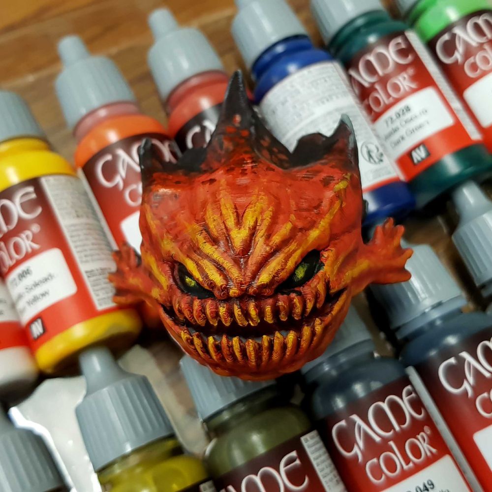 The 3D printed model, now painted. It uses the same colour scheme of yellow red and black as the reference, with some light grey highlights on the sharp edges of the rocky protrusions. The final product is an interpretation of the reference rather than a identical replica. The model is facing the camera, resting on an assortment of small paint bottles. The flying base went back to its owner.