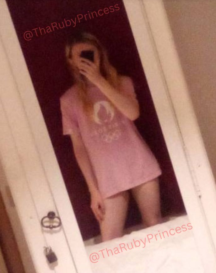 Princess Ruby is wearing a large pink Olympics tshirt, with bare legs, showing bare white thighs, her hand rests on the right. Blonde hair floats down her shoulders and chest and she's posing with her ass slightly curved out in the mirror.