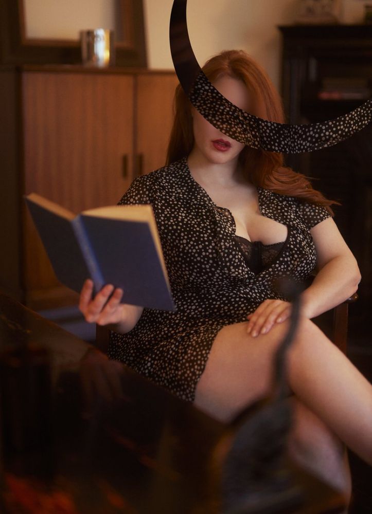 A gorgeous GFE escort Lana van Dam reads a book seated in a chair with her curves on full display. She is visiting NYC from Philly.