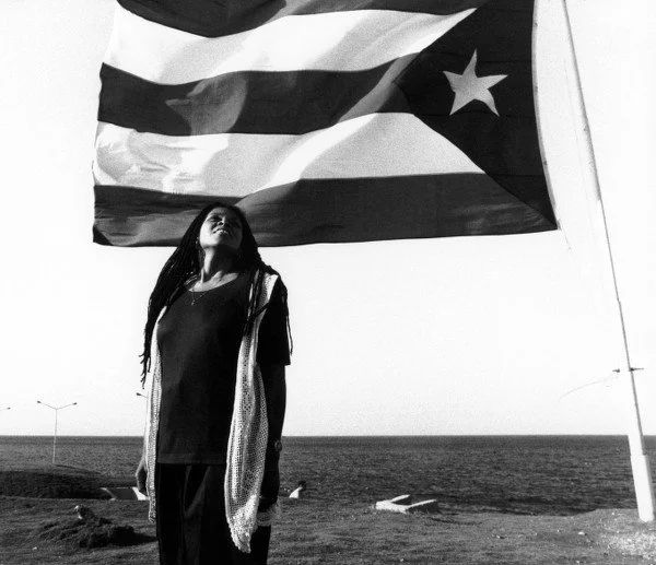 assata standing under a large Cuban flag, black and white photo. from https://www.hamptonthink.org/read/assata-shakur-black-liberation-struggles-and-the-cuban-revolution