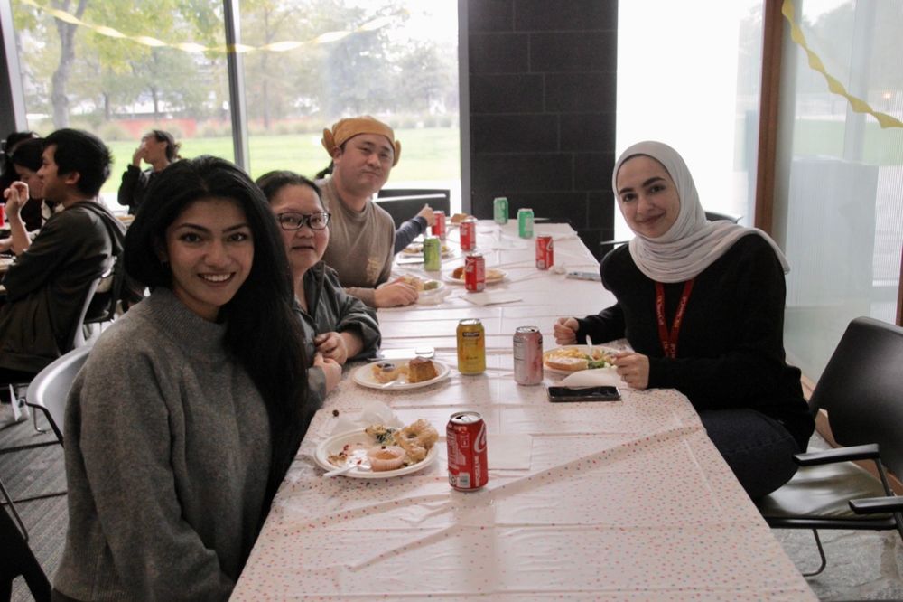 Four people sitting at a table enjoying a meal together in a cafeteria-style setting, with beverages and food plates visible.