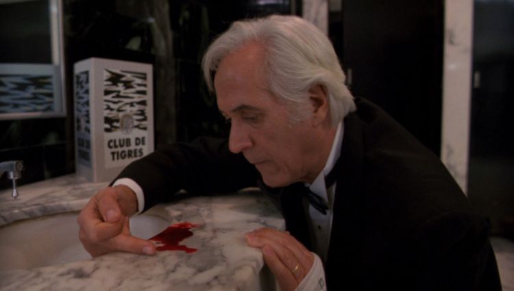 Federico Luppi as Jesús Gris in ‘Cronos’ (1992, dir. Del Toro). Jesús is an older man with white hair wearing a smart black suit with a white shirt. He is kneeling in a bathroom with a wide marble sink. There is a small puddle of blood by the rim. He is curving his hand around the blood.