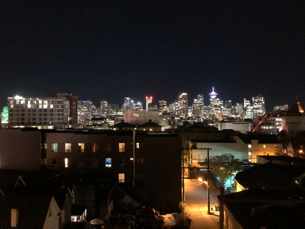 A clear photograph of the same skyline depicting the cityscape of Vancouver, Canada at night with alleyways and apartment buildings in the foreground.